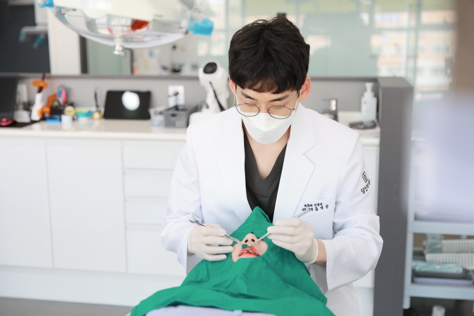 마스크를 착용한 치과의사가 환자의 구강을 진료하는 모습. 환자는 초록색 천으로 얼굴 일부를 가리고 있다.