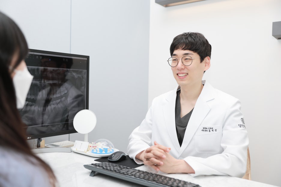 치과의사가 환자와 상담하며 미소 짓는 모습. 진료실 책상에 앉아 컴퓨터 화면을 보고 있다.